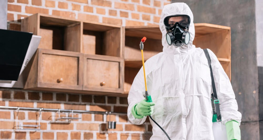 Pest control worker in full protective gear holding a sprayer in a kitchen environment.